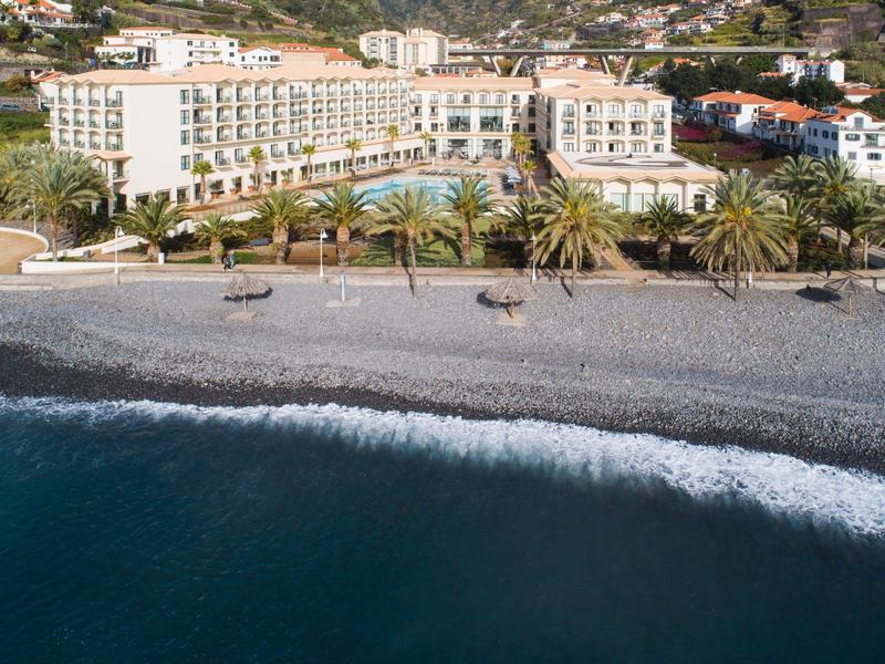 Hotel przy plaży z palmami i ciemną piaszczystą plażą wzdłuż niebieskiej wody oceanu.