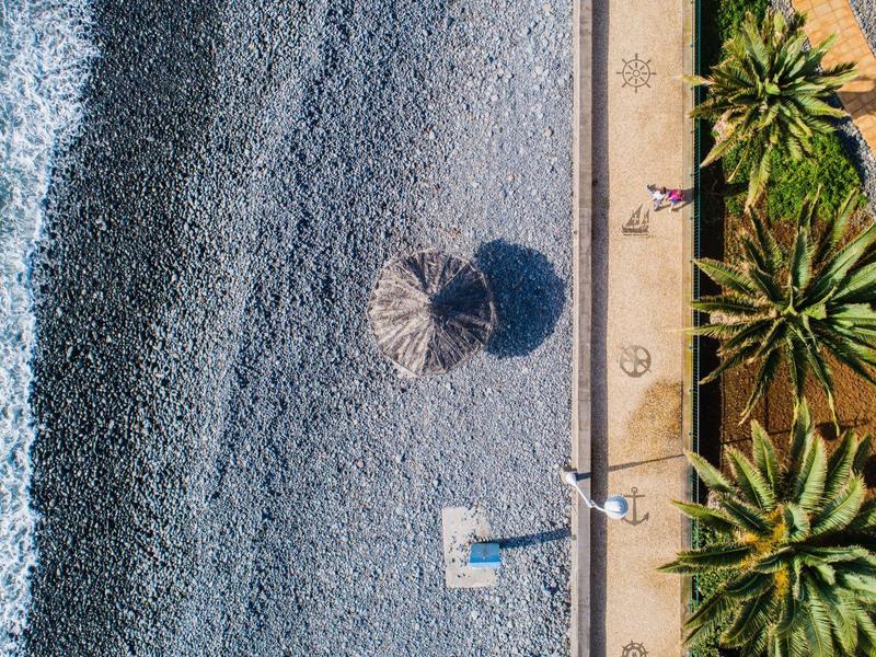 Widok z lotu ptaka na kamienistą plażę z parasolami, palmami i chodnikiem wzdłuż brzegu.