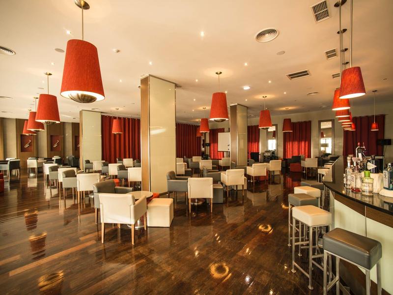 Przestronna restauracja hotelowa z drewnianą podłogą, białymi krzesłami i czerwonymi lampami.