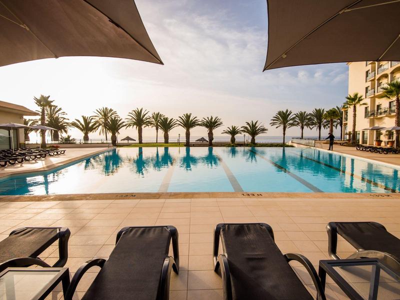 Leżaki przy basenie z widokiem na basen infinity, palmy i parasole o zachodzie słońca.