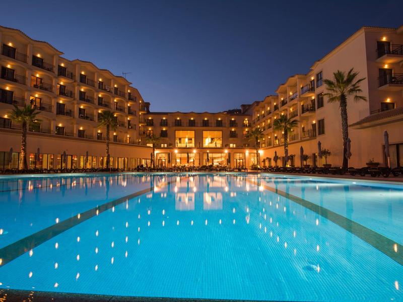 Hotel z oświetlonym basenem o zmierzchu, otoczony balkonami i palmami.