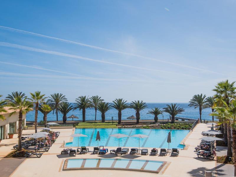 Basen na zewnątrz z leżakami, palmami i widokiem na ocean pod niebieskim niebem.