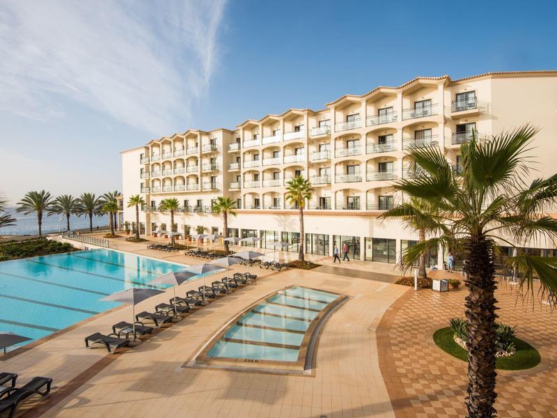 Hotel nad brzegiem morza z basenem, leżakami i palmami pod pochmurnym niebem.