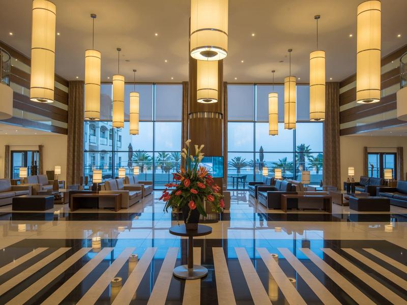 Luksusowy hol hotelowy z wiszącymi lampami, kwiatowym bukietem i dużymi oknami z widokiem na palmy.