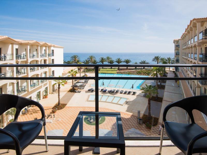 Widok z balkonu na basen, leżaki, palmy i ocean między budynkami hotelu.