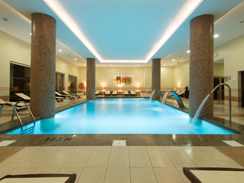 Kryty basen hotelowy z kolumnami i leżakami przy miękkim oświetleniu.