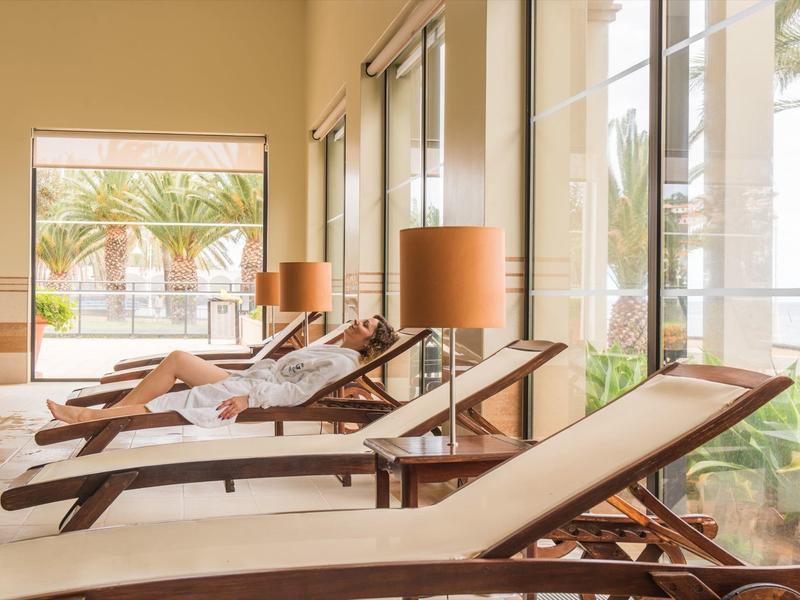 Wewnętrzny salon z drewnianymi leżakami i dużymi oknami z widokiem na palmy na zewnątrz.