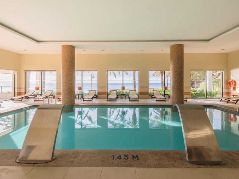 Basen wewnętrzny z dwoma zjeżdżalniami i leżakami przy dużych oknach.