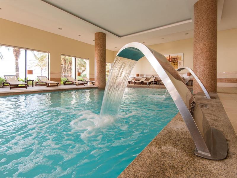 Basen kryty z wodospadem i leżakami przy dużych oknach.