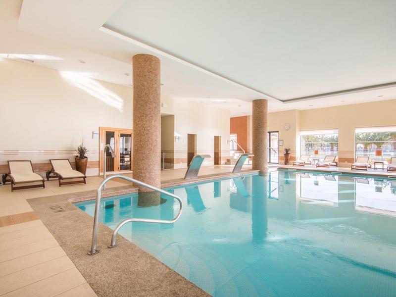 Hotelowy basen kryty z leżakami, filarami i dużymi oknami wpuszczającymi światło naturalne.