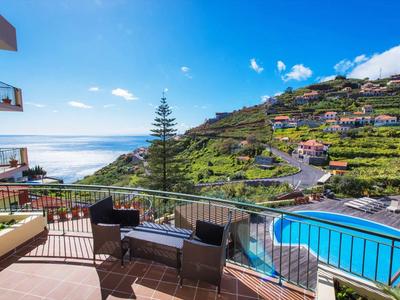 Balcon avec sièges, piscine et vue panoramique sur les collines et la mer sous un ciel clair.