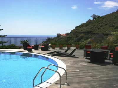 Piscine avec terrasse en bois et chaises longues face à la mer et à la colline en arrière-plan.