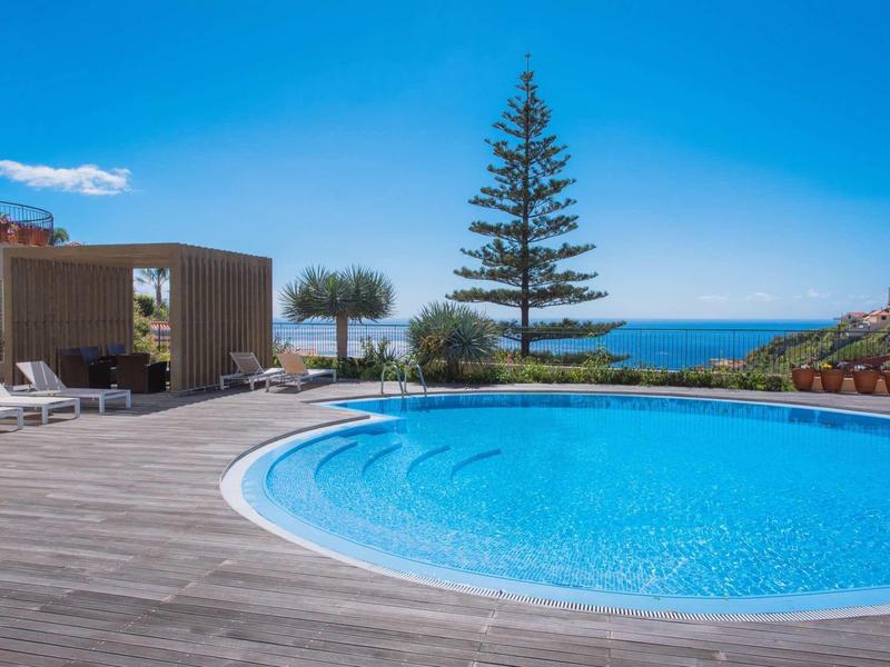 Piscine ronde avec terrasse en bois, chaises longues et vue sur la mer bleue et le ciel dégagé.