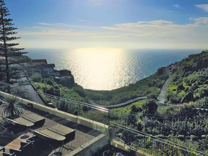 Vue d'une terrasse avec des sièges surplombant la mer et une colline verte au soleil.