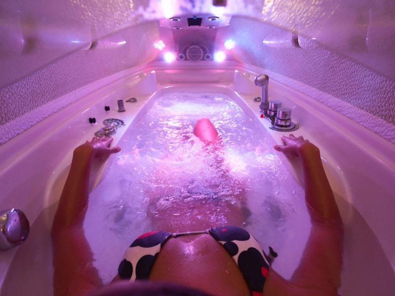 Persona che si rilassa in una vasca idromassaggio con schiuma e illuminazione rosa.