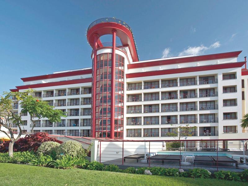 Edificio alberghiero moderno a più piani con torre rossa e grande area erbosa in primo piano.