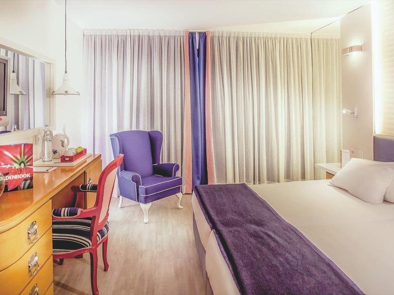 Camera d'hotel luminosa con letto matrimoniale, scrivania, poltrona e grande specchio.