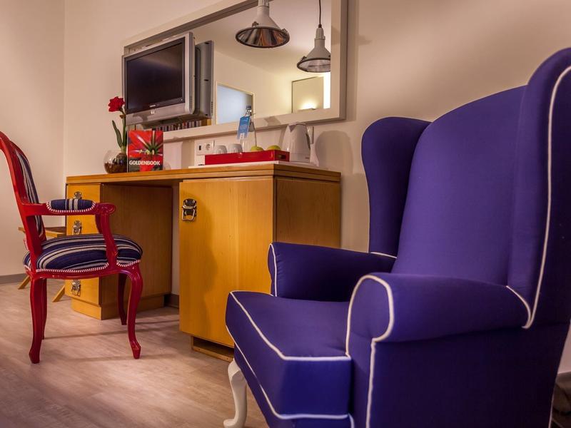 Comoda poltrona blu davanti a una scrivania con sedia rossa e specchio nella camera d'hotel.