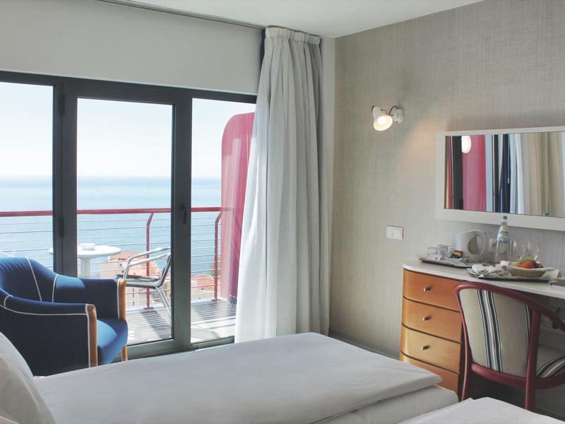 Camera d'hotel con letto, scrivania e vista mare attraverso grandi finestre con balcone