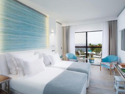 Helles Schlafzimmer mit großem Bett, blauer Wanddekoration, zwei Sesseln und Balkon mit Meerblick.