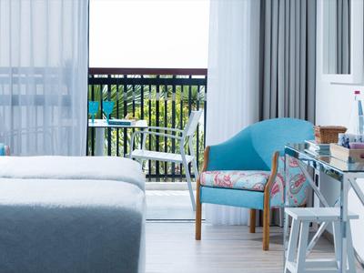 Hell eingerichtetes Zimmer mit blauem Sessel, Glas-Schreibtisch und Blick auf Balkon mit Stühlen.