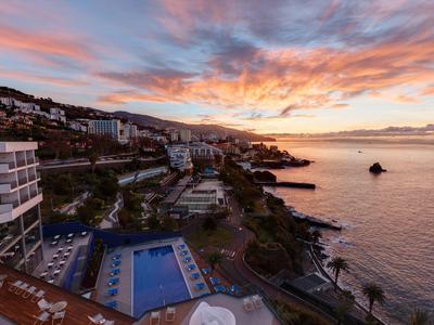 Küstenstadt mit Hotels, Pool, Palmen und farbigem Sonnenuntergang über ruhigem Meer.
