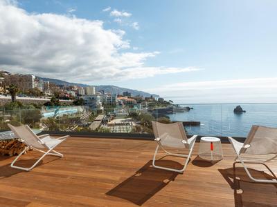 Terrasse mit Holzfußboden, weißen Stühlen und Blick auf Hafen, Meer und bewaldete Hügel im Hintergrund.