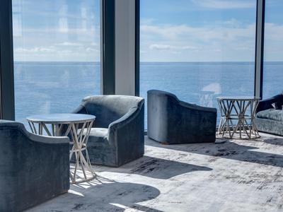 Lounge mit blauen Sesseln und kleinen runden Tischen vor großen Fenstern mit Meerblick.
