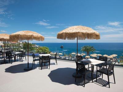 Terrasse mit schwarzen Tischen und Stühlen unter Strohmattenschirmen mit Blick auf das blaue Meer.