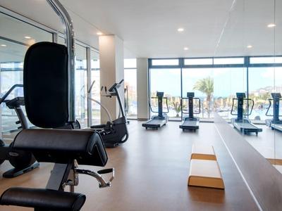 Moderne, helle Fitnessstudio mit Geräten, Spiegelwand und großen Fenstern mit Blick nach draußen.