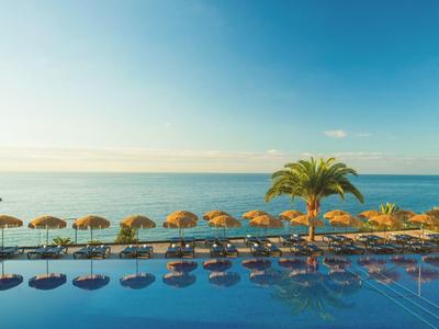 Ein großer Pool mit Sonnenschirmen und Palmen am ruhigen Meer unter blauem Himmel.