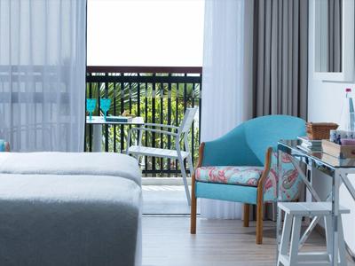 Hell eingerichtetes Schlafzimmer mit blauem Sessel, Glas-Schreibtisch und Blick auf Balkon mit Garten.
