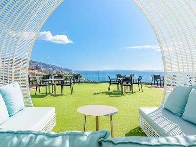 Blick durch weißen Rundpavillon auf grüne Terrasse mit Sitzgruppen und Meer im Hintergrund.