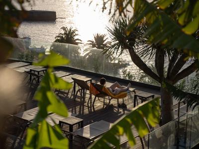Sonnige Terrasse mit leeren Tischen und Stühlen neben einem ruhigen Fluss, umgeben von Pflanzen.
