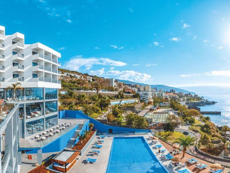 Luxe hotel met zwembaden en terras aan zee onder een blauwe lucht.