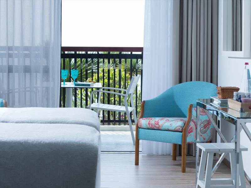 Lichte hotelkamer met bed, blauwe fauteuil en balkon met groen uitzicht.