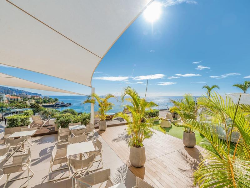 Dakterras met ligstoelen, zonneschermen en helder zeezicht onder fel zonlicht.