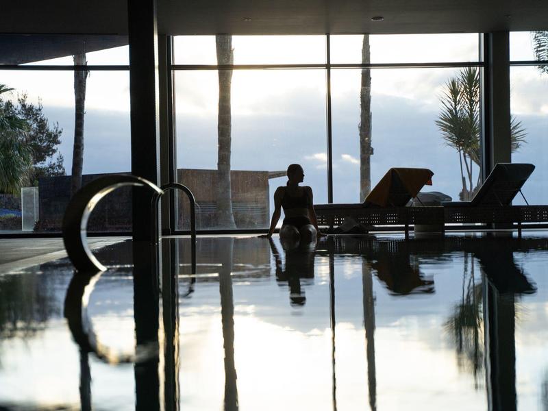 Persoon zit bij een binnenzwembad met ligstoelen en grote glazen ramen op de achtergrond.