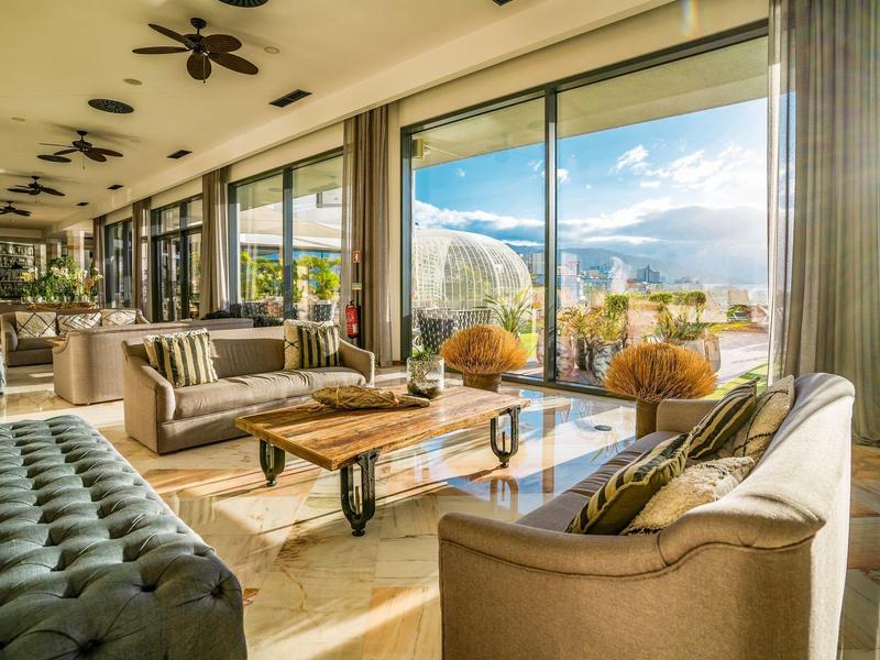 Lichte lounge met grote glazen ramen, banken en uitzicht op zonnig terras en bergen.