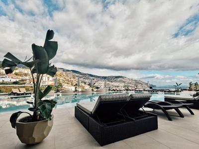 Moderner Poolbereich mit schwarzen Liegen, großer Pflanze im Topf und Blick auf Küstenstadt und Berge.