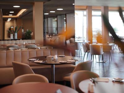 Modernes Restaurant mit hellen Stühlen, runden Tischen und großer Fensterfront im Hintergrund.