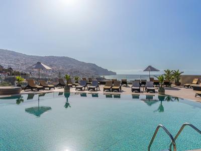 Runder Pool mit Liegen und Sonnenschirmen, dahinter Berge und Meer unter blauem Himmel.