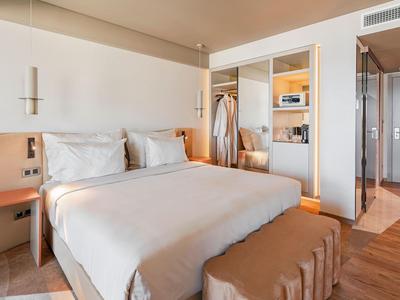 Helles Hotelzimmer mit großem Bett, beigem Fußende, offenem Kleiderschrank und Holzboden.