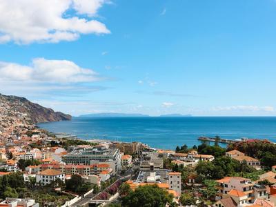 Blick auf eine Küstenstadt mit roten Dächern, Bergen links und blauem Meer unter blauem Himmel.