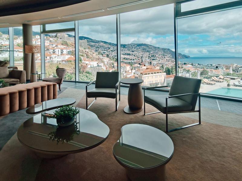 Moderner Wohnraum mit braunem Teppich, Glasfenstern und Blick auf Stadt und Meer bei Tageslicht.