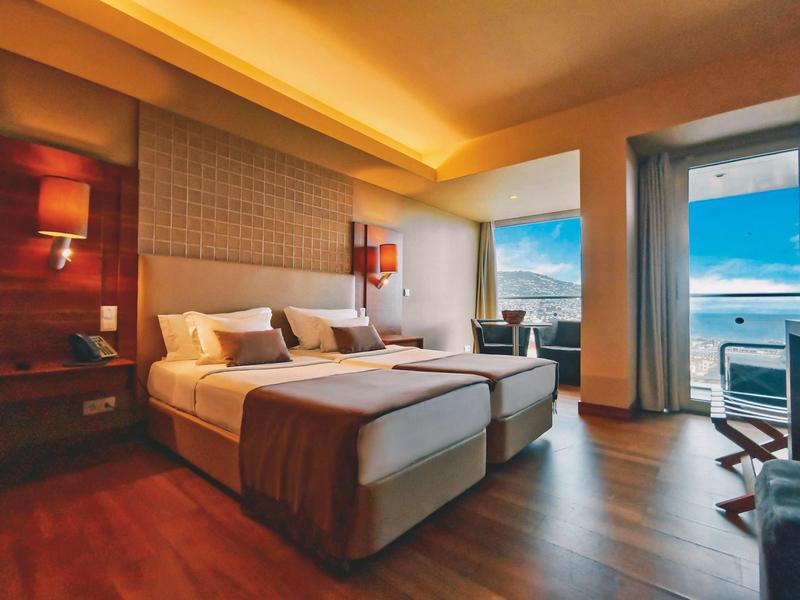 Modernes Schlafzimmer mit zwei Betten, Holzboden, warmen Wänden und Blick auf Meer durch große Fenster.