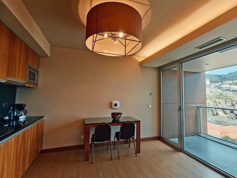 Moderner Raum mit Holzmöbeln, dunklem Tisch, zwei Stühlen, großer Fensterfront und Deckenlampe.