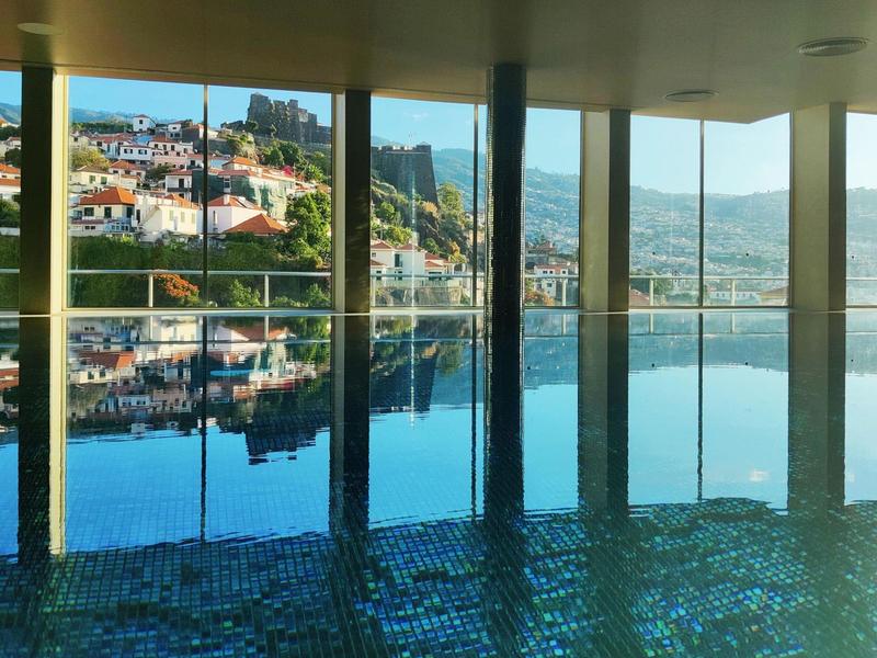 Hallenbad mit Glasfront, Blick auf Stadt und Hügel, Spiegelung im Wasser, blaue Fliesen am Boden.