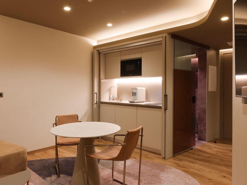 Moderner Wohnraum mit rundem weißen Tisch, zwei Holzstühlen und integrierter Küchenzeile in warmem Licht.