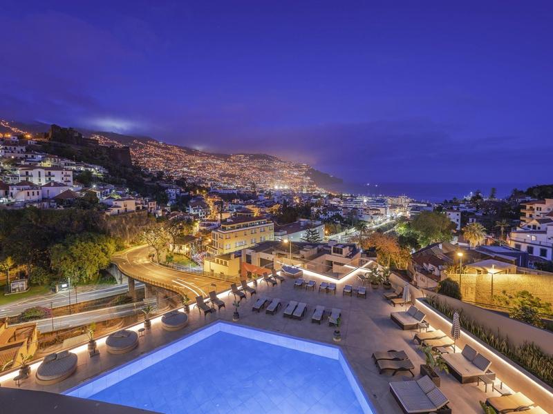 Nächtliche Stadtansicht mit beleuchtetem Pool und umliegenden Gebäuden in hügeliger Landschaft.